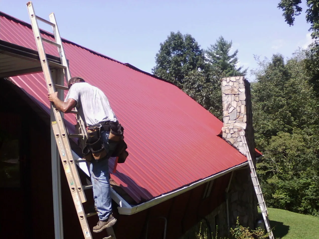 Roofer on a ladder beside a red metal roof and stone chimney — Soffit & Fascia Repair by Roofing Brothers in Whitakers